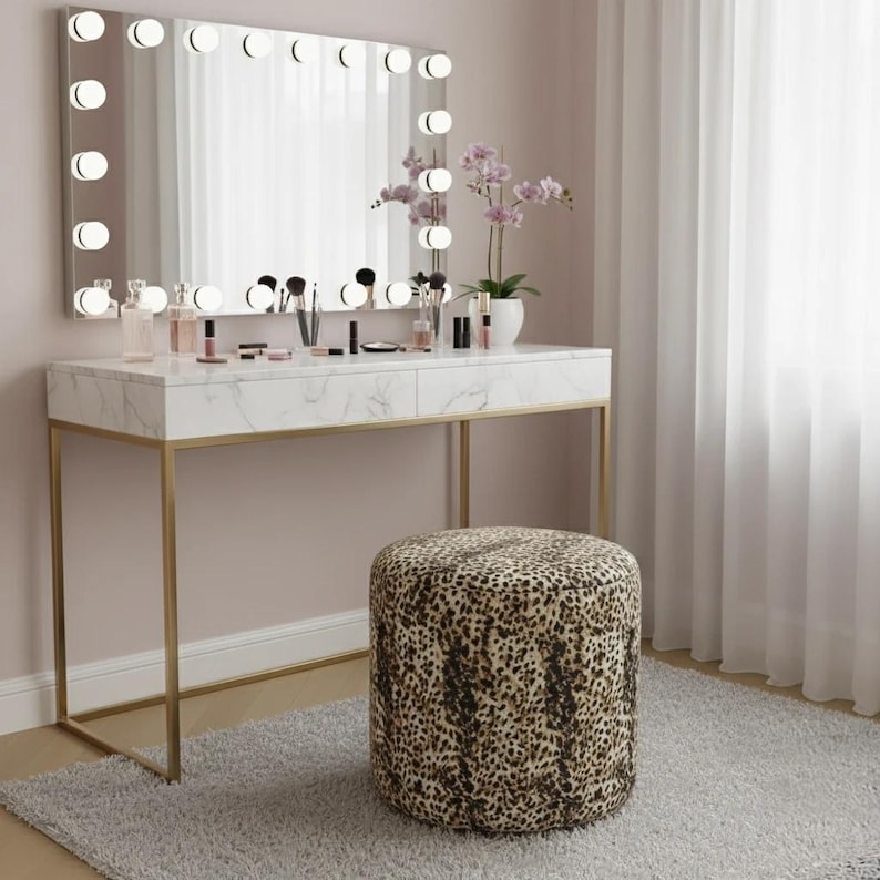May include: A vanity set with a marble-look top, gold legs, and a large mirror with lights. The set includes a leopard-print stool, makeup, and a potted orchid. The room has a neutral color palette with a light gray rug and white curtains.