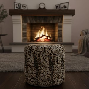 May include: A round ottoman with a leopard print pattern. The ottoman is brown and tan with a dark brown spotted design. The background includes a fireplace with a burning fire, a rug, and a dark wood floor.