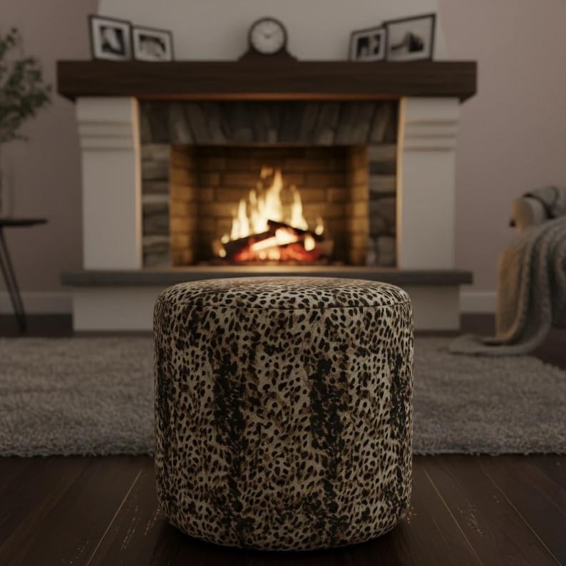 May include: A round ottoman with a leopard print pattern. The ottoman is brown and tan with a dark brown spotted design. The background includes a fireplace with a burning fire, a rug, and a dark wood floor.