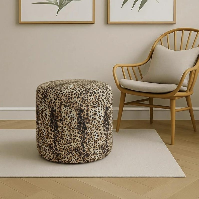 May include: A cylindrical ottoman with a leopard print pattern. The ottoman is brown, tan, and black. It is placed on a light beige rug in front of a wooden chair with a beige cushion. Two framed botanical prints hang on the wall.