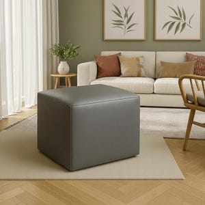 May include: A square, gray ottoman in a living room setting. The ottoman is made of a smooth material and sits on a neutral-colored rug. A white sofa with decorative pillows is in the background.