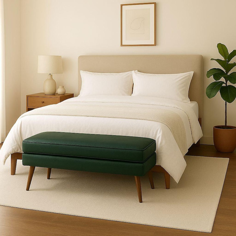 May include: A bedroom scene featuring a bed with white bedding, pillows, and a beige headboard. A dark green bench with wooden legs sits at the foot of the bed. A beige rug and a potted plant complete the room's decor.