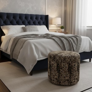 May include: A bedroom scene featuring a navy blue tufted headboard bed with white bedding and a gray throw blanket. A leopard print ottoman sits in front of the bed on a light gray rug. A lamp and side table are visible in the background.