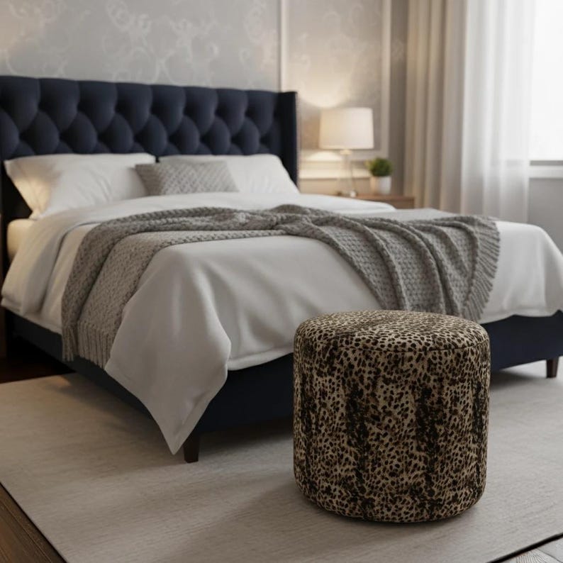 May include: A bedroom scene featuring a navy blue tufted headboard bed with white bedding and a gray throw blanket. A leopard print ottoman sits in front of the bed on a light gray rug. A lamp and side table are visible in the background.