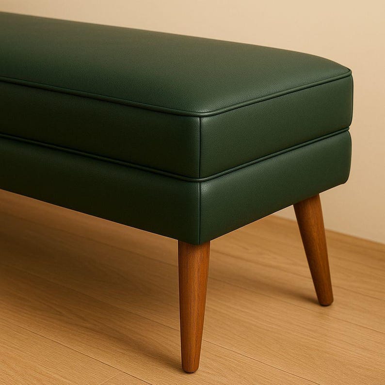 May include: A dark green bench with a rectangular seat and wooden legs. The bench has a stitched border and a mid-century modern design. The legs are angled and made of light brown wood.