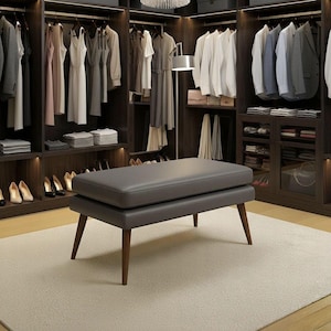 Pode incluir: Um banco de couro cinza com pernas de madeira está em um closet. O closet apresenta prateleiras de madeira escura e roupas penduradas, incluindo vestidos, camisas e jaquetas. Sapatos estão cuidadosamente organizados nas prateleiras, e uma luminária de chão está por perto.