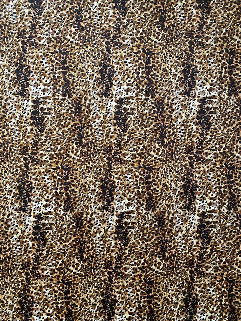May include: A close-up of a fabric with a leopard print pattern. The pattern features a mix of brown, black, and tan spots on a cream-colored background. The spots vary in size and shape, creating a textured appearance.