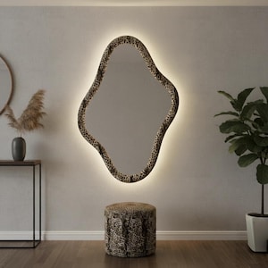 May include: A large, uniquely shaped mirror with a leopard print frame and a soft, glowing light around the edges. A matching leopard print ottoman sits below the mirror. The room has a neutral color palette with a plant and decorative vase.