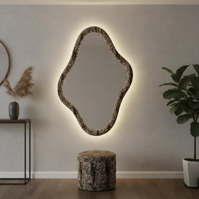 May include: A large, uniquely shaped mirror with a leopard print frame and a soft, glowing light around the edges. A matching leopard print ottoman sits below the mirror. The room has a neutral color palette with a plant and decorative vase.