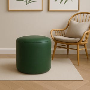 Pode incluir: Um pufe cilíndrico verde esmeralda. O assento redondo e estofado é feito de um material liso. O pufe está sobre um tapete bege claro, com uma cadeira de madeira e obras de arte emolduradas ao fundo.