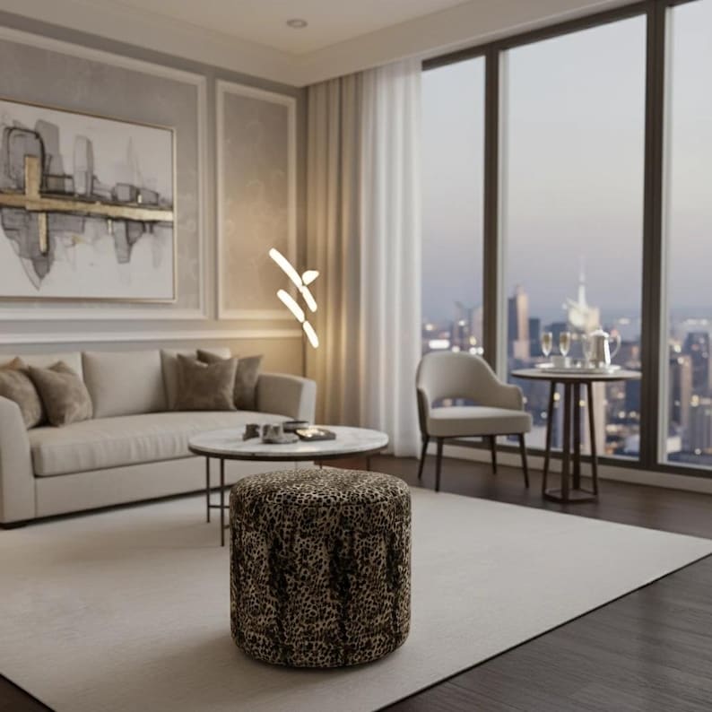 May include: A modern living room with a neutral color palette. A leopard-print ottoman sits on a light-colored rug. A sofa with throw pillows, a round coffee table, and a side table with a chair are also present. Large windows offer a city view.