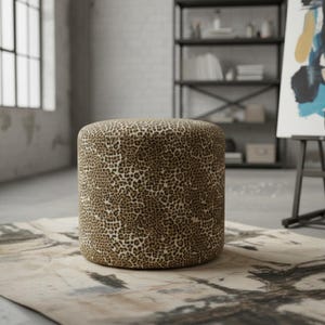 May include: A cylindrical ottoman with a leopard print pattern. The brown and tan spotted fabric covers the entire surface of the footstool. The ottoman is placed on a patterned rug in a room with a window and a metal shelf.