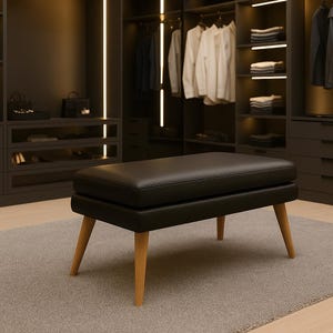 Pode incluir: Um pufe de couro preto com um tampo retangular e quatro pernas de madeira clara anguladas. O pufe está colocado sobre um tapete cinza num closet, com prateleiras de madeira escura e roupas visíveis ao fundo.
