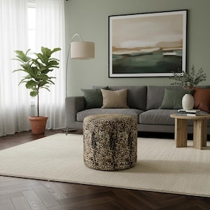 May include: A living room scene featuring a gray sofa with decorative pillows, a round ottoman with a leopard print, and a potted plant. A large framed artwork hangs above the sofa, and a beige rug covers the floor.