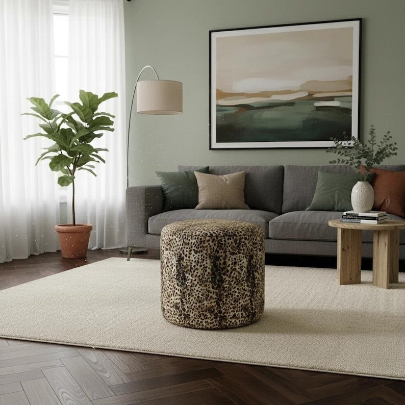 May include: A living room scene featuring a gray sofa with decorative pillows, a round ottoman with a leopard print, and a potted plant. A large framed artwork hangs above the sofa, and a beige rug covers the floor.