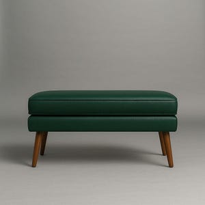 May include: A dark green upholstered ottoman with a rectangular shape. The ottoman has a padded top and sits on four wooden legs. The legs are angled and have a natural wood finish. The background is a solid gray.