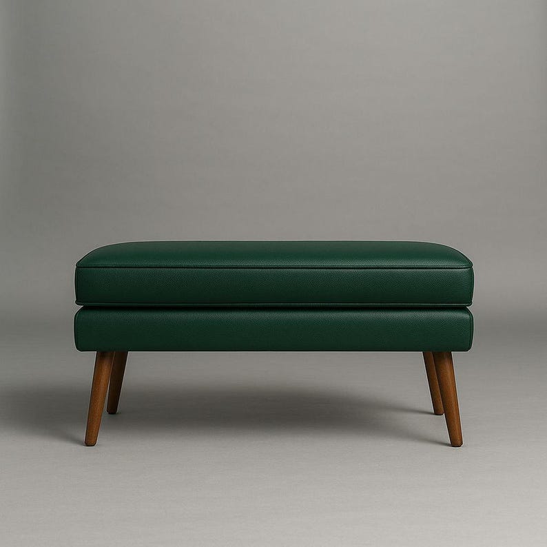 May include: A dark green upholstered ottoman with a rectangular shape. The ottoman has a padded top and sits on four wooden legs. The legs are angled and have a natural wood finish. The background is a solid gray.
