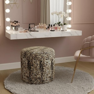 May include: A vanity set with a leopard print ottoman, a marble-look floating shelf, and a pink chair. The shelf holds makeup, brushes, and a mirror. The room has a soft, feminine aesthetic with a large mirror and warm lighting.