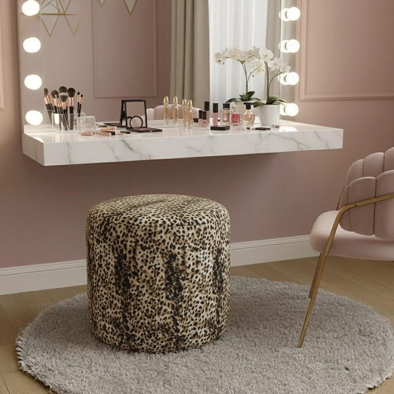 May include: A vanity set with a leopard print ottoman, a marble-look floating shelf, and a pink chair. The shelf holds makeup, brushes, and a mirror. The room has a soft, feminine aesthetic with a large mirror and warm lighting.