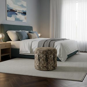 May include: A bedroom scene featuring a teal upholstered bed with white bedding and decorative pillows. A round ottoman with a leopard print sits in front of the bed. A painting and sheer curtains are visible.
