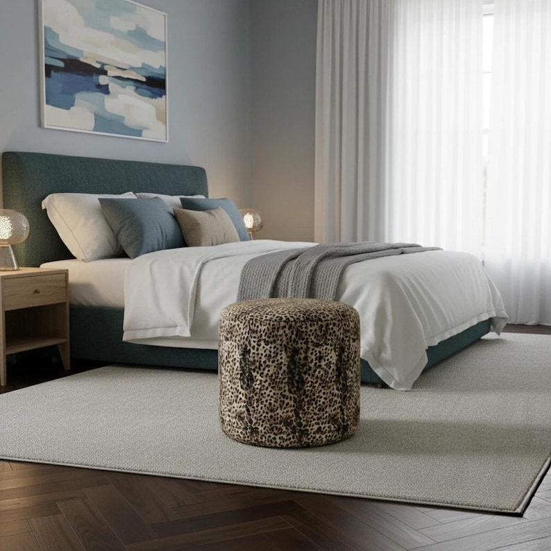 May include: A bedroom scene featuring a teal upholstered bed with white bedding and decorative pillows. A round ottoman with a leopard print sits in front of the bed. A painting and sheer curtains are visible.