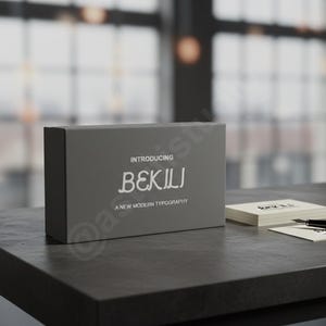 May include: A gray box with the text "INTRODUCING BEKILI A NEW MODERN TYPOGRAPHY" sits on a dark gray table. Beside it are business cards and a black pen. The background is blurred, with a window and soft lighting.