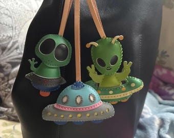 May include: Three leather alien-themed keychains. Two green aliens with large black eyes and a light green body. One alien is in a blue and pink UFO. The keychains are attached to a leather strap. The background is a black bag.
