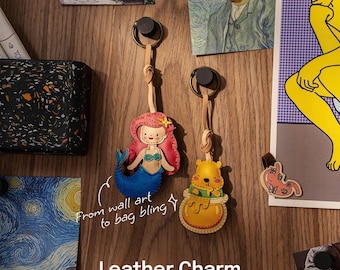 May include: Two leather charms hang from black magnetic clasps. One charm is a mermaid with a red tail and pink hair. The other is a yellow bear in a honey pot. The text "Leather Charm" is at the bottom.