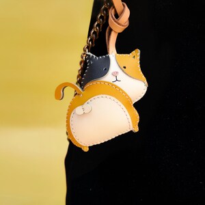 May include: A small, leather cat-shaped keychain with a black, white, and yellow color scheme. The keychain is attached to a brown leather strap and a gold chain.