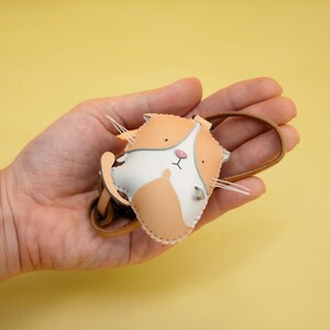 May include: A small, hand-stitched leather cat keychain with a white face and orange fur. The keychain is held in a hand against a yellow background.