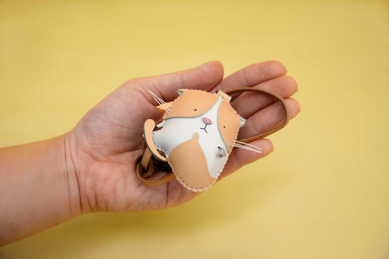 May include: A small, hand-stitched leather cat keychain with a white face and orange fur. The keychain is held in a hand against a yellow background.