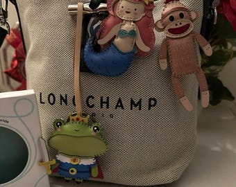 May include: A canvas tote bag with the word "LONGCHAMP" printed on it. Three decorative charms hang from the bag: a mermaid with pink hair and a blue tail, a sock monkey, and a frog wearing a crown and holding a sword.