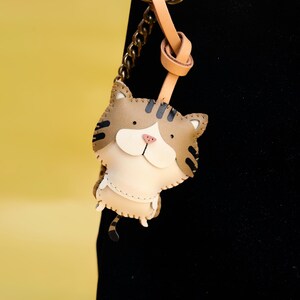 May include: A brown and white leather cat keychain with black stripes. The keychain is attached to a brown leather strap and a gold chain.