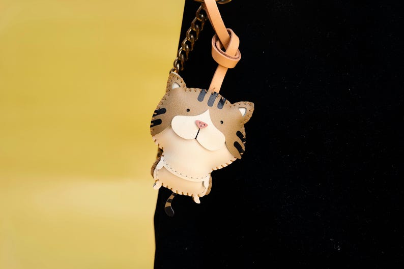 May include: A brown and white leather cat keychain with black stripes. The keychain is attached to a brown leather strap and a gold chain.