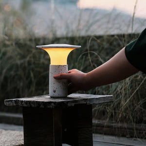 May include: A white speckled table lamp with a round, flat top and a warm, glowing light. The lamp is sitting on a wooden stool.