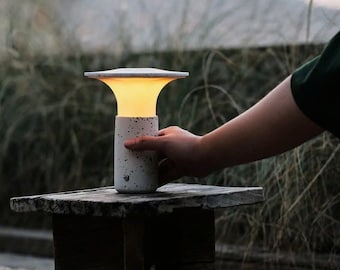 May include: A white speckled table lamp with a round, flat top and a warm, glowing light. The lamp is sitting on a wooden stool.