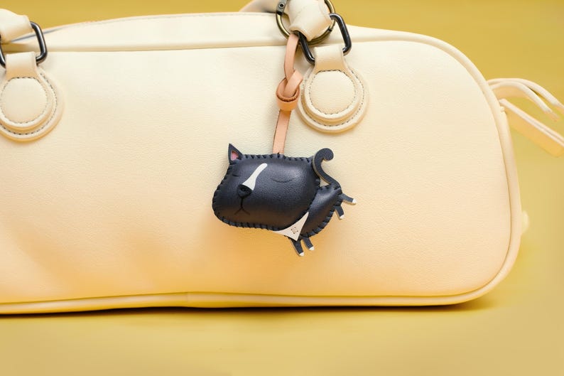 May include: A cream-colored leather handbag with a black leather cat charm attached to the handle. The cat charm has white accents on its face and a black collar.