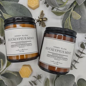 May include: Two glass jars of Eucalyptus Mint beeswax candles with black lids and white labels. The labels read "Honey Glow Eucalyptus Mint". The image includes a gold bee figurine and two honeycomb-shaped wax melts. The candles are 6 oz / 250g.