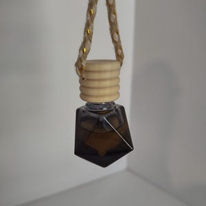 May include: A hanging car air freshener with a geometric, dark brown glass bottle. The bottle is topped with a light wood cap and a braided rope with gold accents. The air freshener is suspended against a white background.