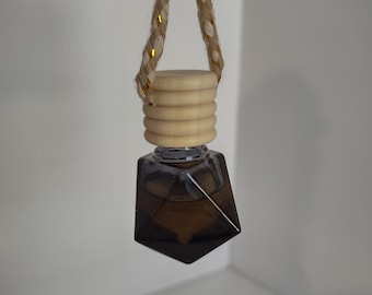 May include: A hanging car air freshener with a geometric, dark brown glass bottle. The bottle is topped with a light wood cap and a braided rope with gold accents. The air freshener is suspended against a white background.