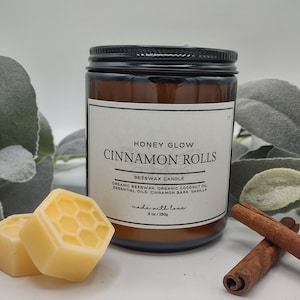 May include: A brown glass jar candle with a black lid, labeled "HONEY GLOW CINNAMON ROLLS." The label lists ingredients and "8 oz / 250g." Beside the candle are two honeycomb-shaped wax melts and two cinnamon sticks. The candle is a beeswax candle.