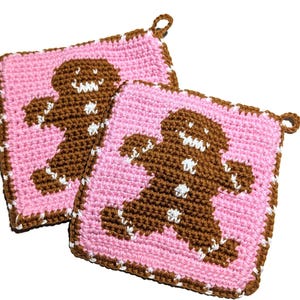 May include: Two square crochet potholders featuring a brown gingerbread man figure centered on a light pink background. Each potholder has a brown border with white stitching details and a small loop at the corner for hanging in the kitchen.