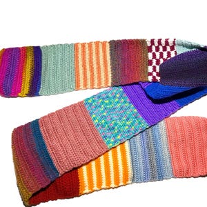 Puede incluir: Una bufanda larga y rectangular de ganchillo compuesta por varios paneles multicolores. La bufanda presenta secciones con colores sólidos, rayas verticales y patrones moteados en tonos rosa, naranja, azul, morado, rojo y verde, unidos entre sí.
