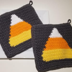 Peut inclure: Deux maniques carrées au crochet en fil gris foncé, chacune présentant un motif de bonbon maïs centré. Le motif est composé de segments de fil blanc, orange et jaune. Chaque manique comprend une petite boucle dans le coin supérieur.