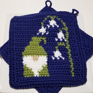Puede incluir: Un agarrador de cocina cuadrado de ganchillo azul oscuro que presenta un gnomo verde con barba blanca y un sombrero alto y puntiagudo. Junto al gnomo hay un tallo verde con flores de lirio de los valles blancas en forma de campana y una pequeña asa.