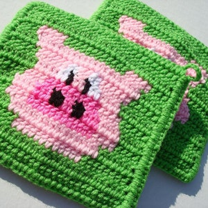 May include: Two handmade crochet potholders. Each potholder features a pink pig face on a green background. The pig has black eyes and nostrils. The potholders are square and have a loop for hanging.