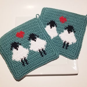 May include: Two square teal crochet potholders featuring a repeating pattern of two sheep with black heads and white bodies, accompanied by a small red heart above them. Each potholder includes a small loop at the corner for convenient hanging.