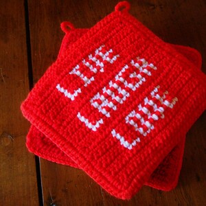 May include: Two red crocheted potholders with the words "LIVE LAUGH LOVE" stitched in white. The potholders are square with a loop for hanging. The potholders are on a wooden surface.