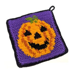 Könnte beinhalten: Ein quadratischer Häkel-Topflappen mit einem zentralen orangefarbenen Kürbismotiv mit schwarzem Gesicht und grünem Stiel auf violettem Hintergrund. Das gesamte Stück ist schwarz umrandet und hat eine kleine schwarze Aufhängeschlaufe an der Ecke.