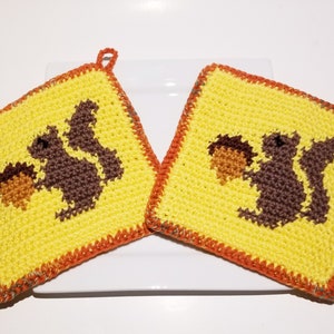Könnte beinhalten: Zwei gelbe gehäkelte Topflappen mit einem braunen Eichhörnchen, das eine Eichel hält. Die Topflappen haben einen orangefarbenen Rand und eine Schlaufe zum Aufhängen. Die quadratischen Topflappen liegen auf einer weißen Oberfläche.